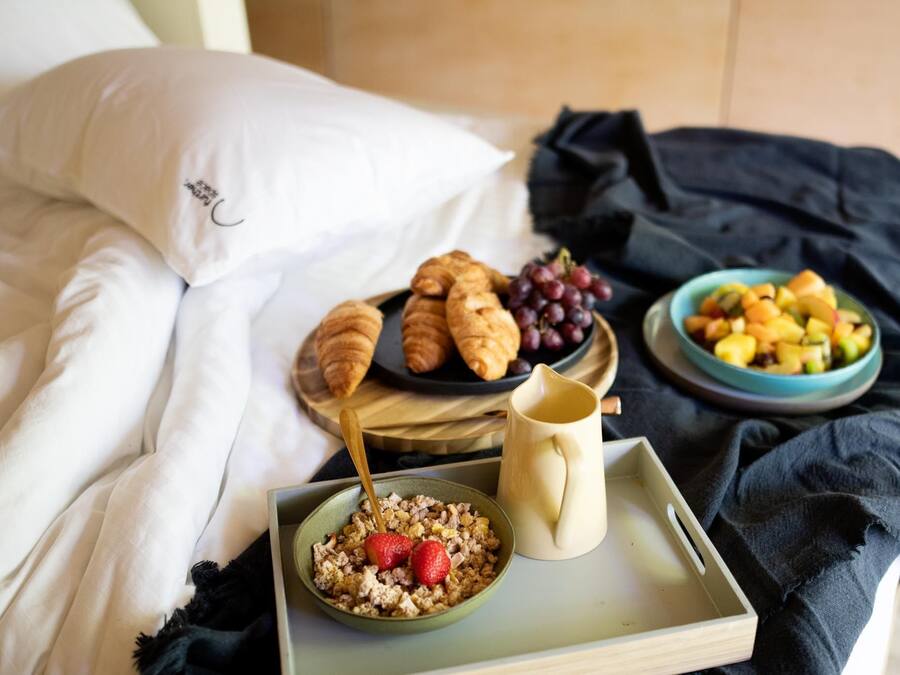 A bed with white linens, a bowl of cereal with strawberries, a jug, and a tray with croissants, grapes, and a fruit bowl.
