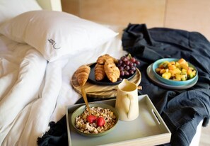 A bed with white linens, a bowl of cereal with strawberries, a jug, and a tray with croissants, grapes, and a fruit bowl.
