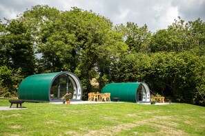 Two green, semi-cylindrical structures with glass doors, surrounded by a grassy area with a wooden table and chairs, and a bench.