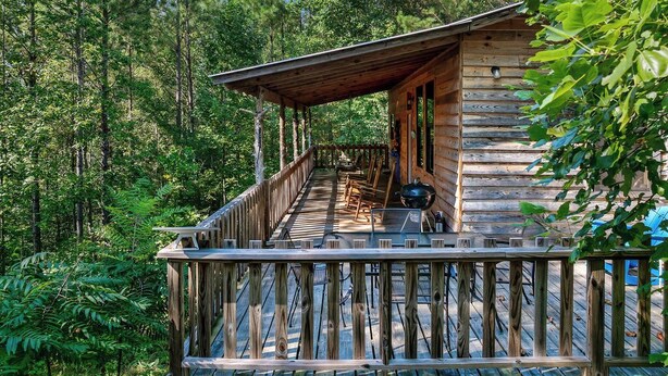 A rustic cabin with a wooden deck, surrounded by dense forest.