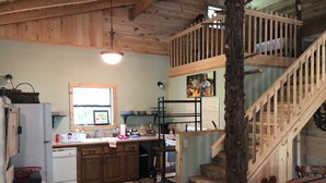 A rustic kitchen with wooden cabinets, a sink, and a window. There is a staircase with a wooden railing leading to an upper level.