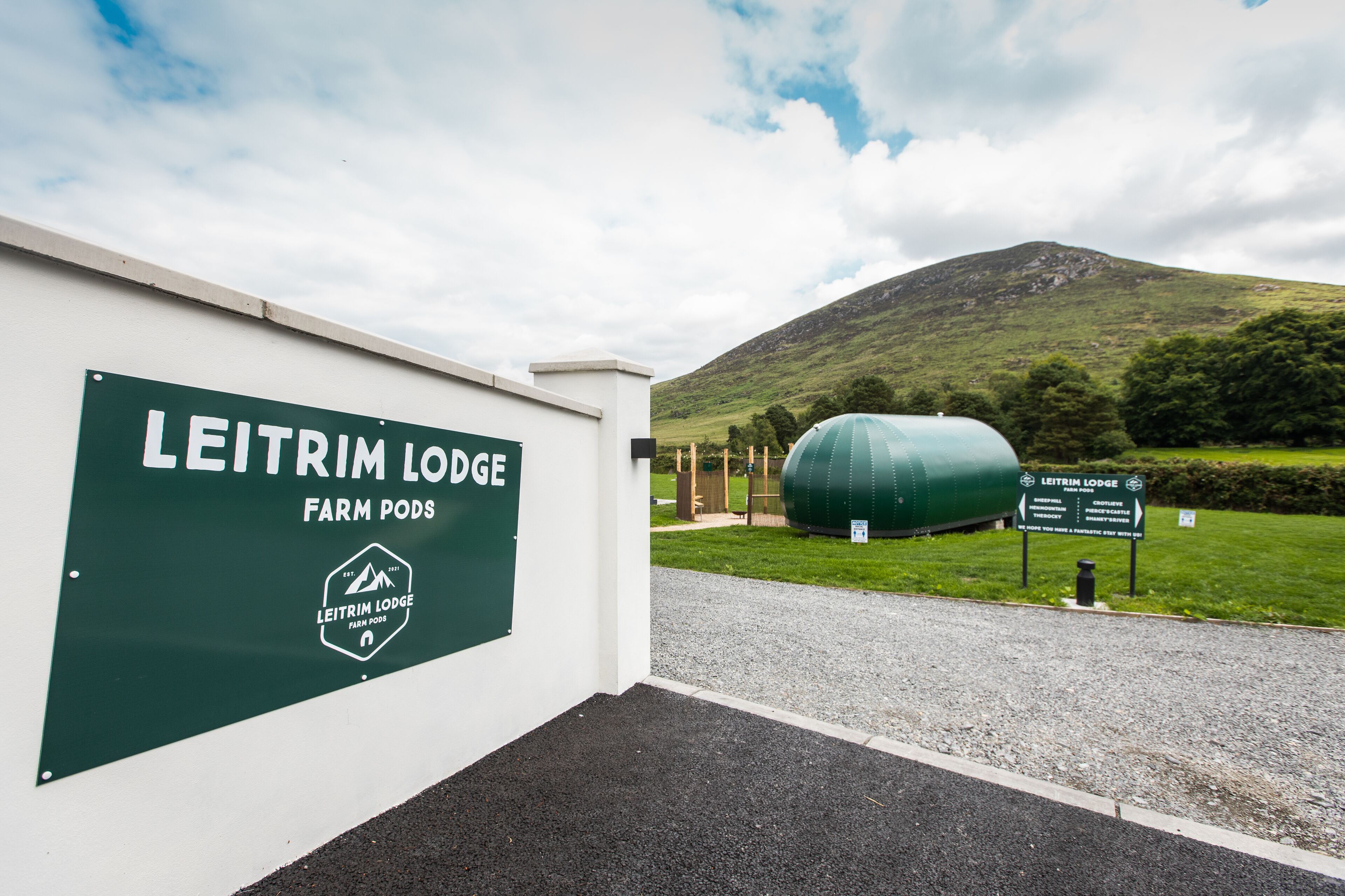 A sign for Leitrim Lodge Farm Pods with a view of a hill and a green tank in the background.