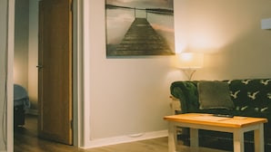 A room with a wooden floor, a green sofa, a small wooden table, a painting of a dock, and a ceiling light fixture. - The Black Isle Apartment (Fortrose)