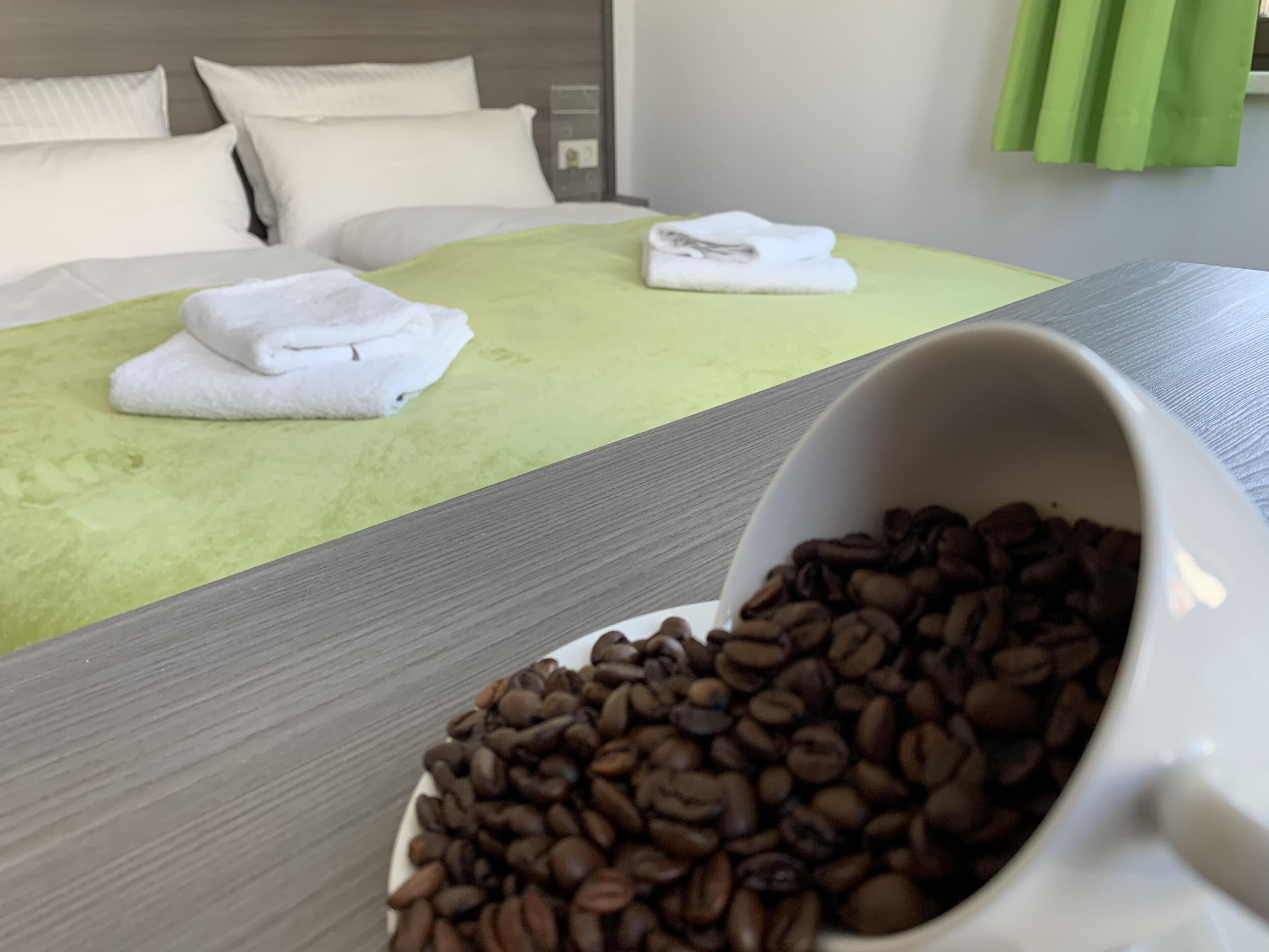 A hotel room with a bed, green bedspread, white pillows, and a coffee cup on the table.