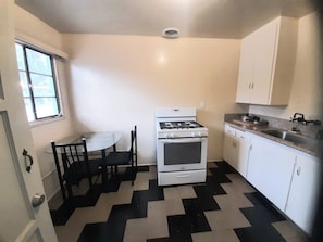 A small kitchen with a gas stove, oven, white cabinets, a dining table with chairs, and a window. - The New Star Motel Experience (Moreno Valley)
