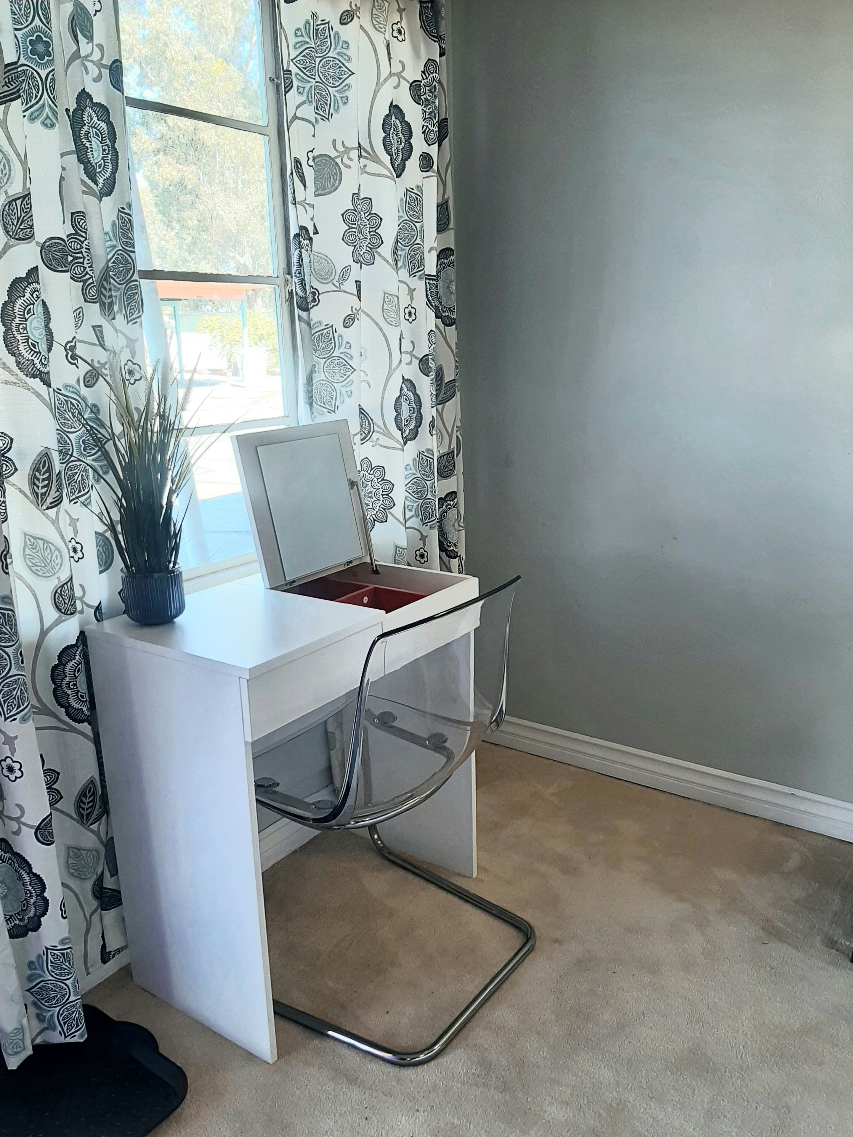 A white desk with a laptop, a potted plant, and a clear chair in a room with patterned curtains and a grey wall.