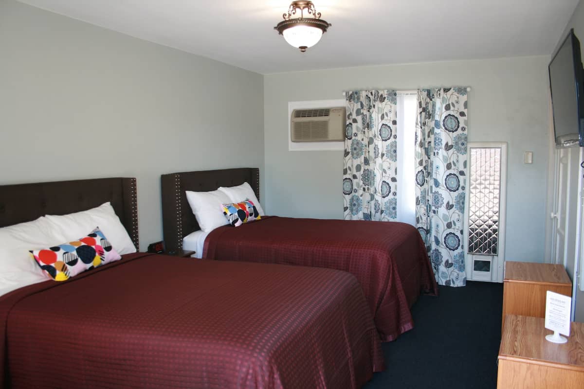A hotel room with two beds, a ceiling light, an air conditioning unit, patterned curtains, and a wooden desk with a sign.
