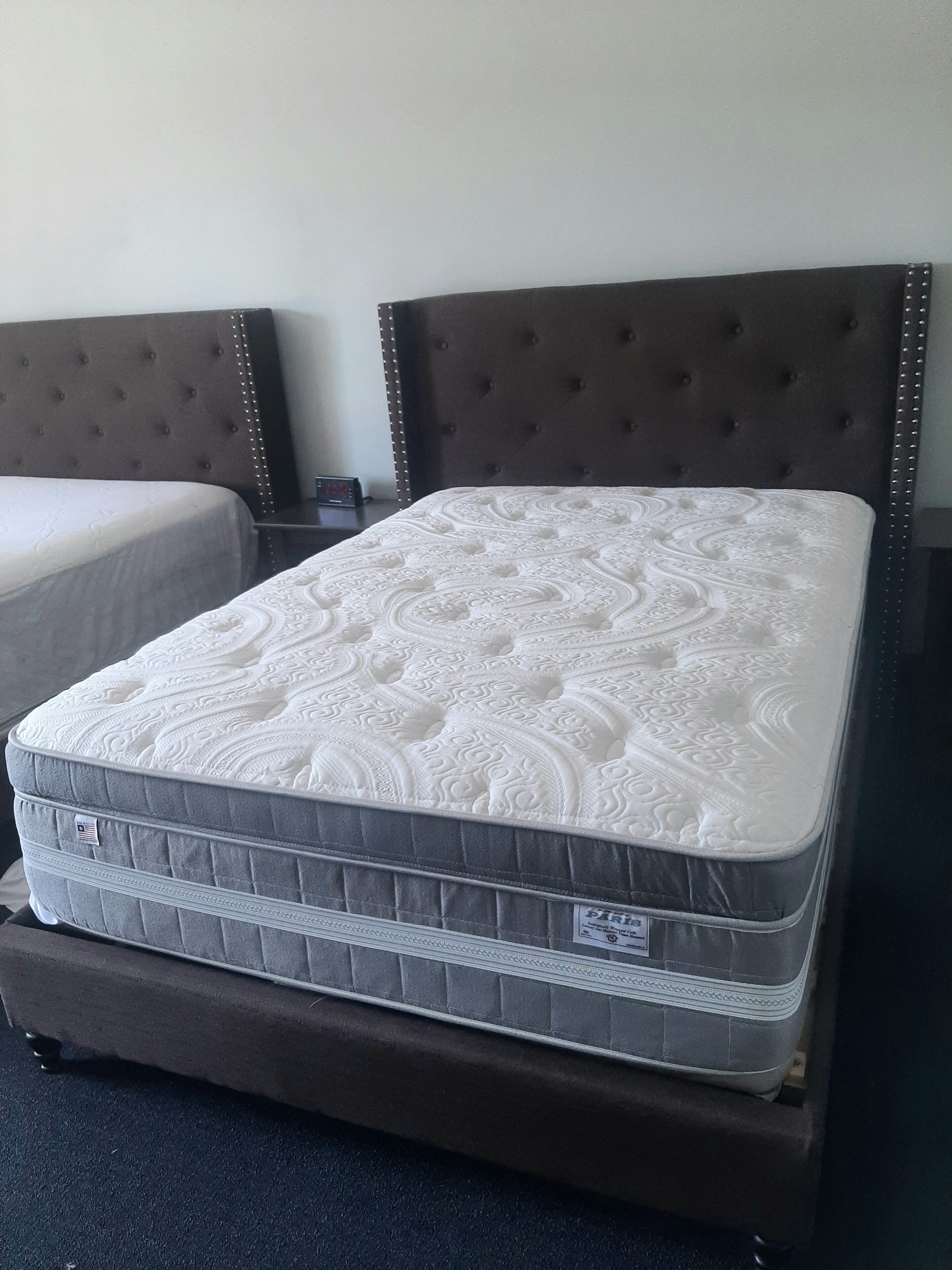 Un llit amb un capçal amb borla i un matalàs estampat en una habitació amb una paret senzilla i un petit rellotge a la nit.