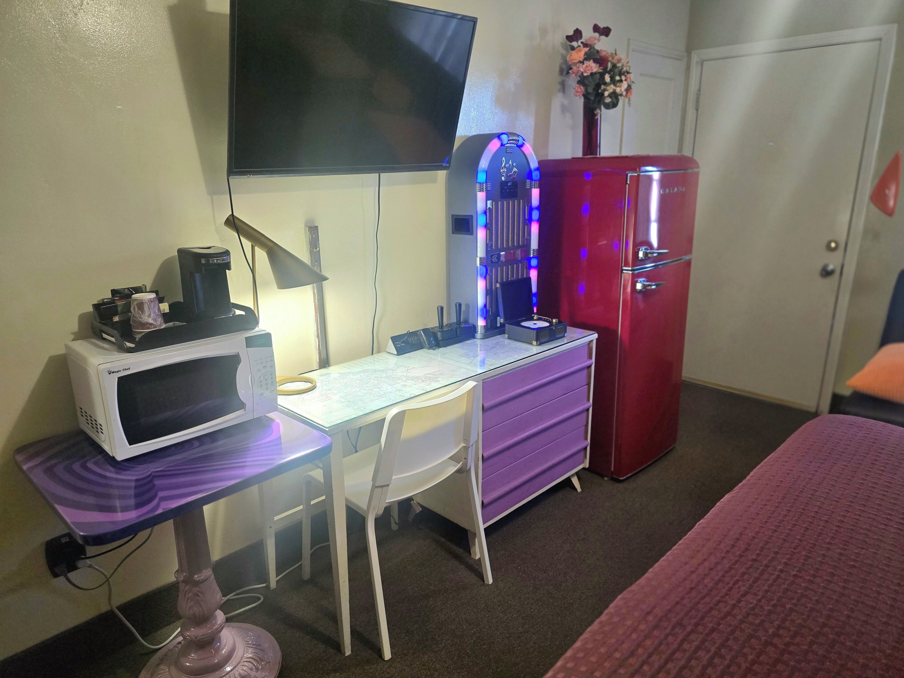 Une chambre avec un bureau, un téléviseur à écran plat, un réfrigérateur rouge, une lampe rose et un lit.