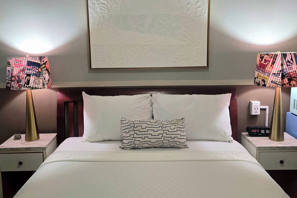 A hotel room with a large bed, two nightstands with lamps, a wall clock, and a framed floral artwork above the bed.