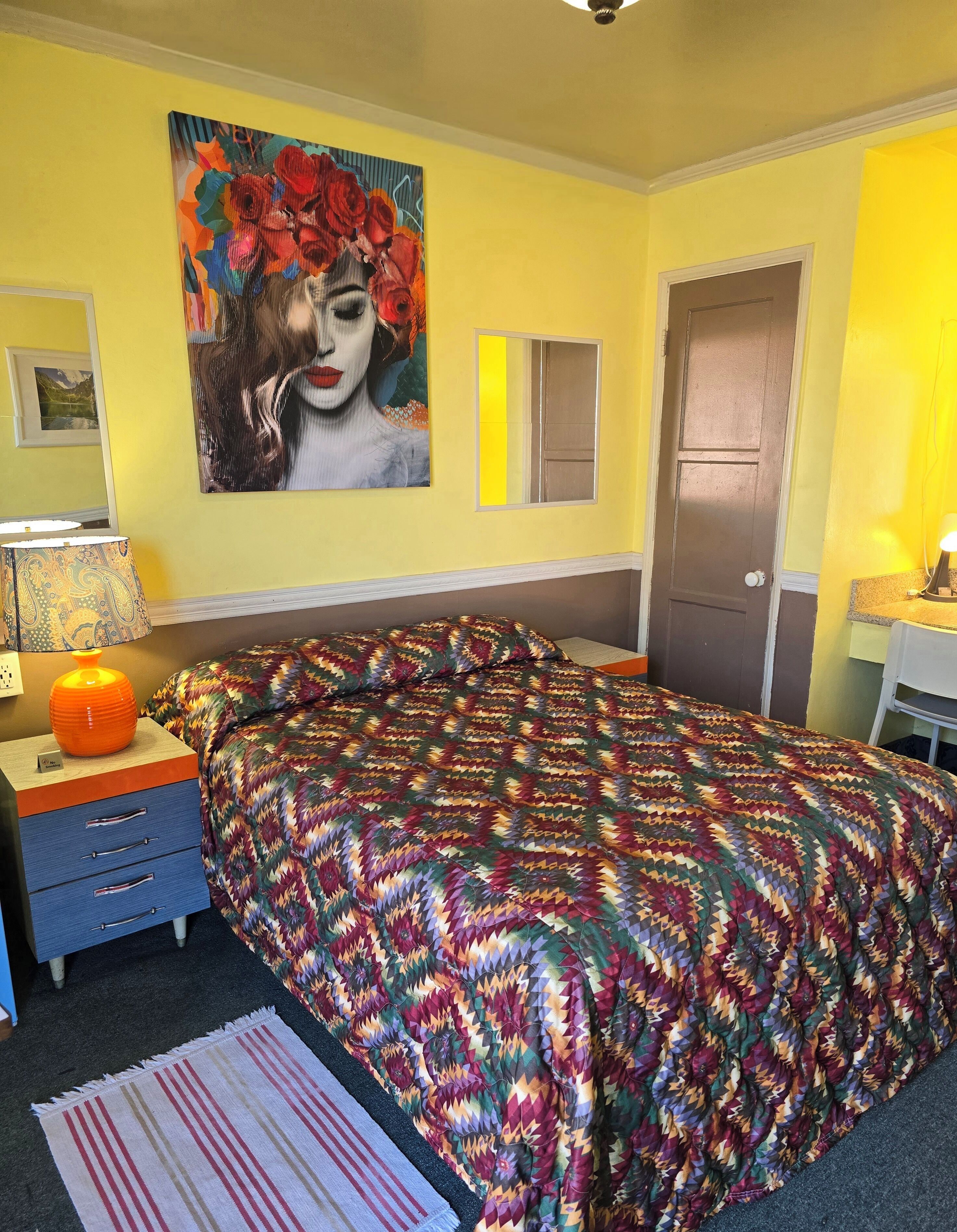 A bedroom with a bed, a nightstand, a lamp, a door, and a painting on the wall.