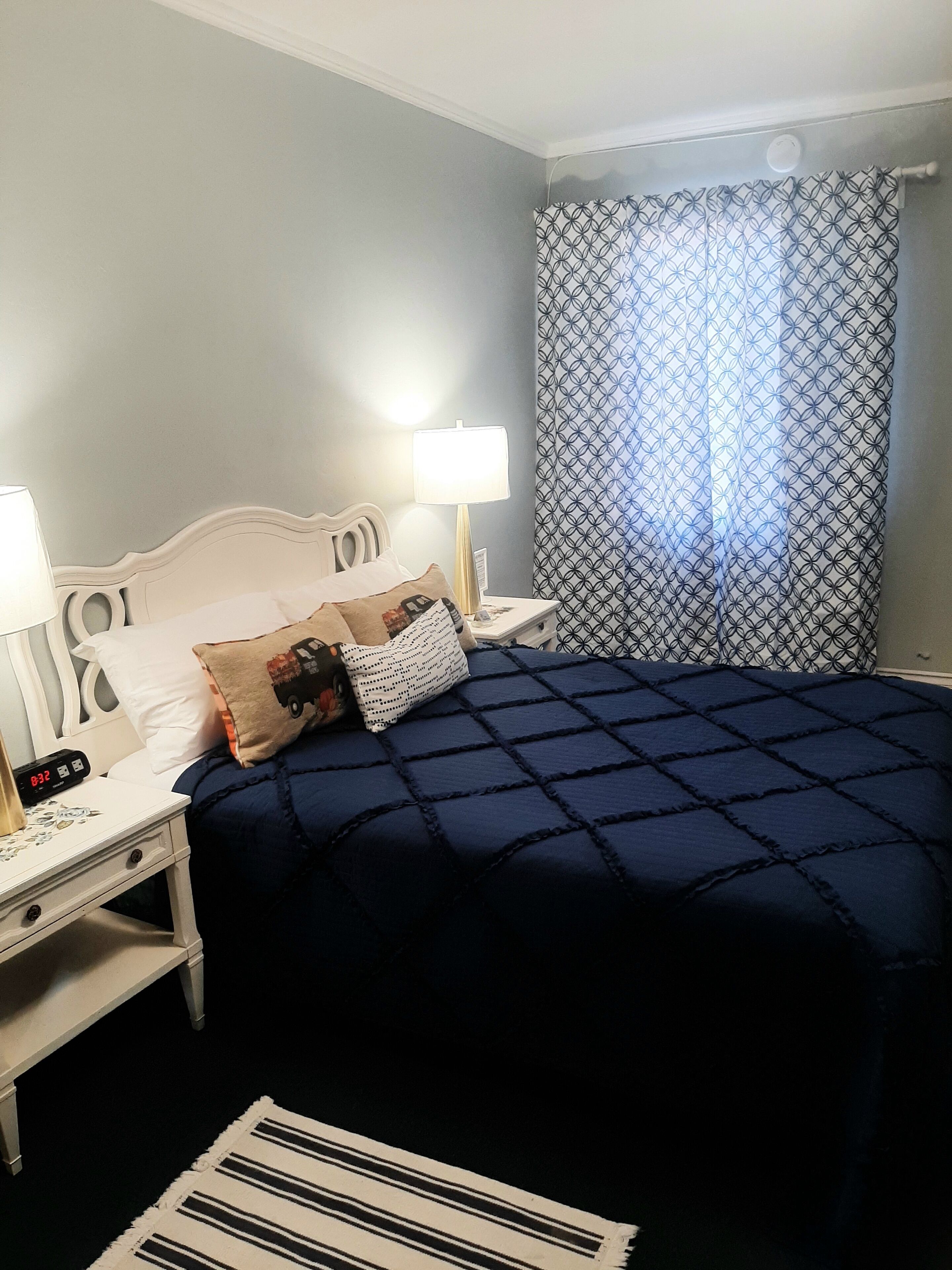 A neatly made bed with a blue quilt, a bedside lamp, a clock, and a patterned curtain.