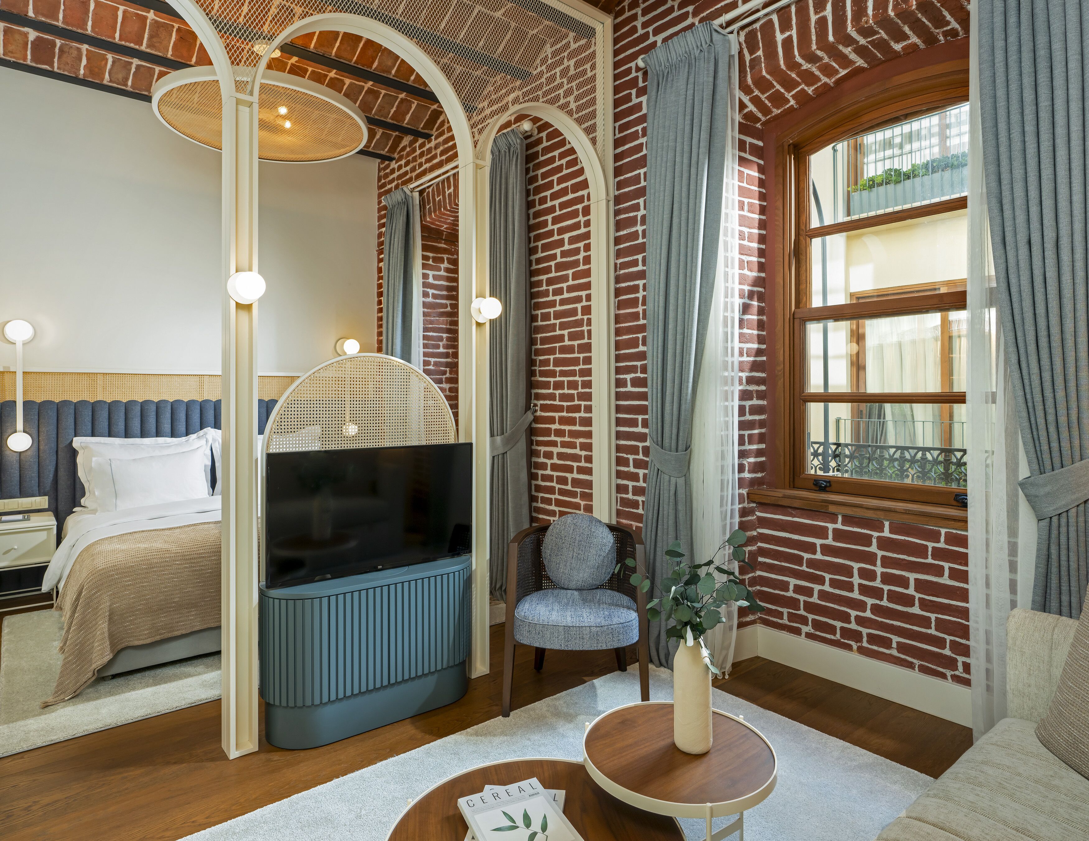 A modern bedroom with a brick wall, a large window with curtains, a bed with a TV, and a small table with a vase.