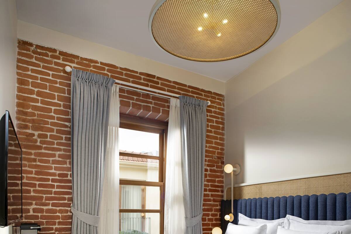 A hotel room with a brick wall, a large window with curtains, a bed with white bedding, a bedside table with a lamp, and a chair with a woven seat.