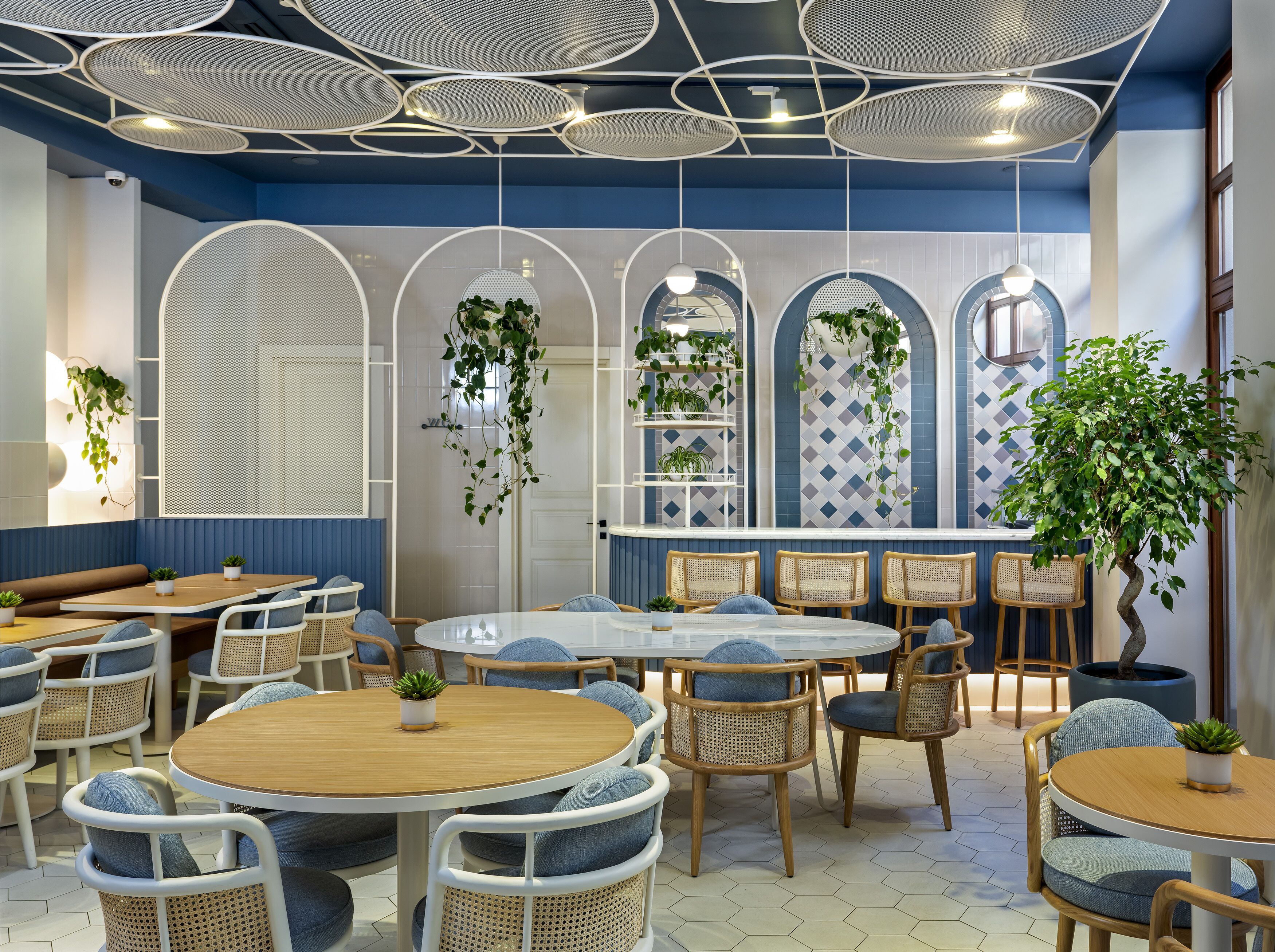 Een modern café met ronde tafels en rieten stoelen, met een blauw-wit kleurenschema en hangplanten.