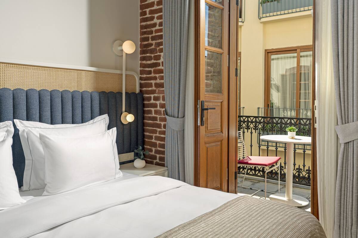 A hotel room with a bed, a nightstand, a lamp, a balcony with a table and chair, and a view of the outside.