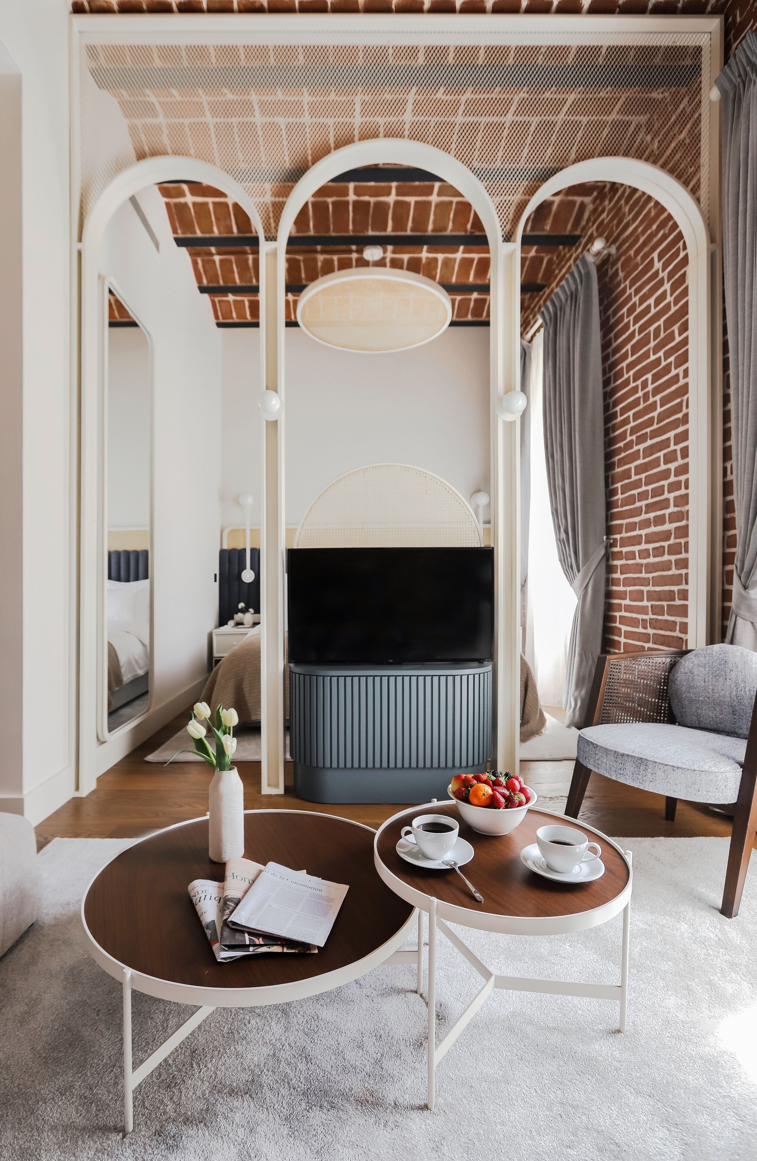 Une chambre d’hôtel moderne avec un mur en briques, un grand miroir, un téléviseur à écran plat, un fauteuil confortable de couleur grise et une petite table ronde sur laquelle se trouvent un saladier rempli de fruits et des tasses.
