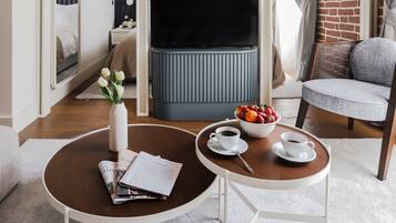 Une chambre d’hôtel moderne avec un mur en briques, un grand miroir, un téléviseur à écran plat, un fauteuil confortable de couleur grise et une petite table ronde sur laquelle se trouvent un saladier rempli de fruits et des tasses.