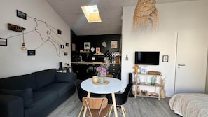 A compact living space with a dark-colored sofa, a round dining table with chairs, a kitchenette, a television, and a door.