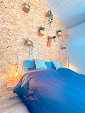 Quarto com parede de madeira, cama com roupa de cama azul e vários vasos suspensos.