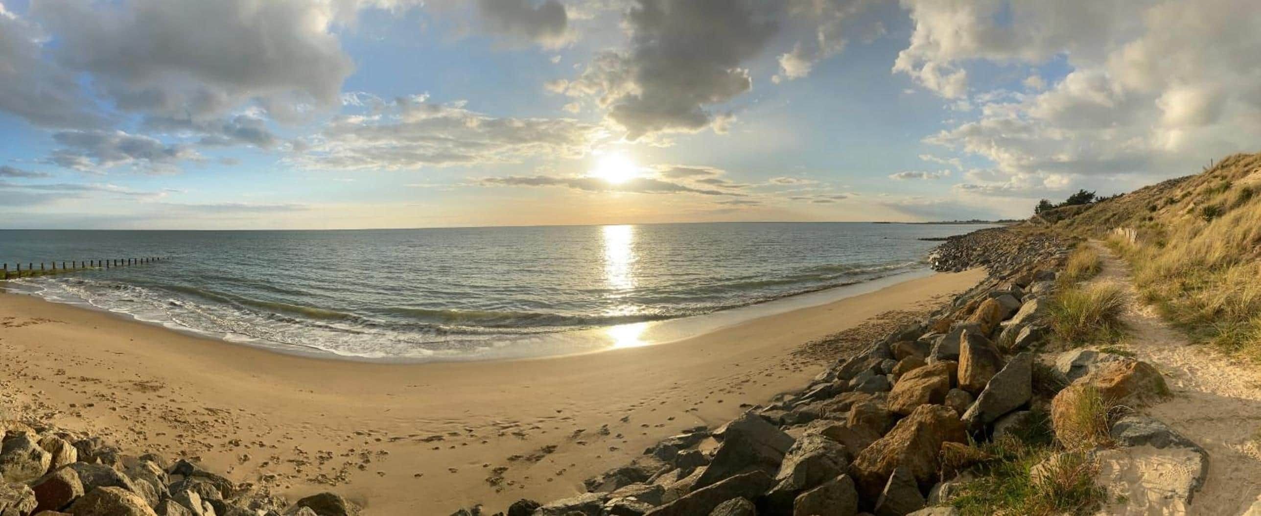 Une plage bordée d’un rivage rocheux, un ciel dégagé et le soleil se couchant à l’horizon.