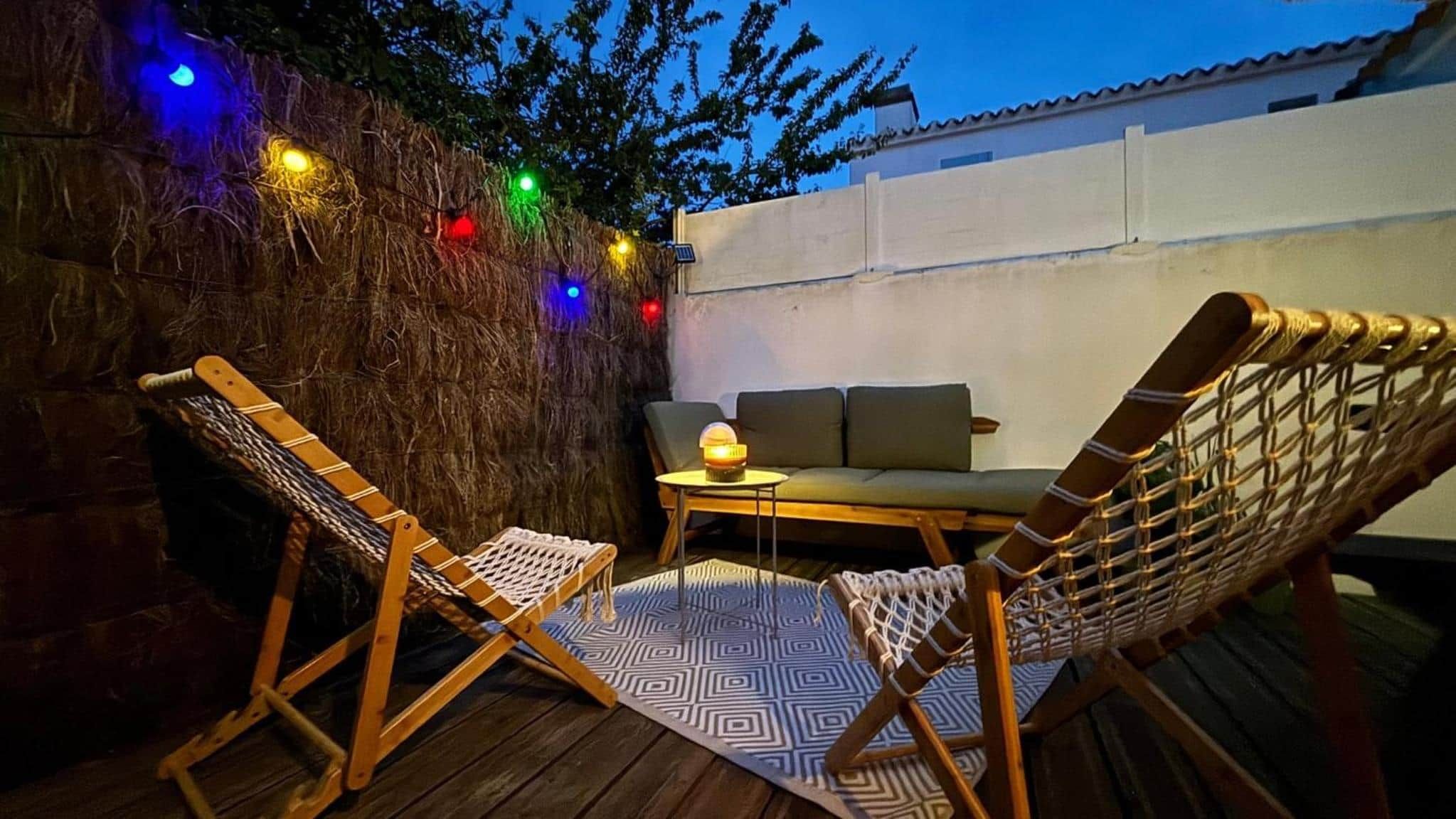 Un espace extérieur aménagé avec deux chaises en bois et une table, entouré d’un mur décoré de guirlandes lumineuses colorées.