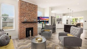 A modern living room with a brick fireplace, comfortable seating, a flat-screen TV, and a kitchen in the background.