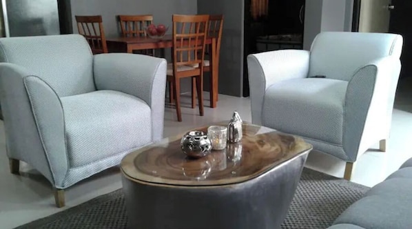 Un salon moderne avec deux fauteuils à accoudoirs recouverts d’un tissu gris, une table basse en bois agrémentée d’objets décoratifs, et un coin repas à l’arrière-plan.