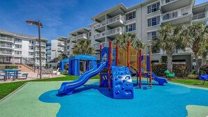 Un patio de juegos con toboganes y estructuras para escalar, rodeado por un edificio con balcones y palmeras.