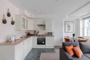 Een moderne keuken met witte kasten, een magnetron en een fornuis. Er is een eethoek met een tafel en stoelen, en een grijze bank met oranje kussens.