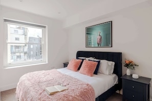 A bedroom with a bed, pillows, a bedside table, a window, and a framed picture on the wall.