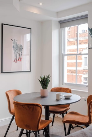 A dining area with a round table, four orange chairs, a framed zebra artwork, a potted plant, and a window with a view of buildings.