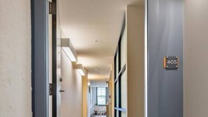 Un couloir d’hôtel avec un paillasson, une porte portant un numéro et une vue sur une chambre avec une table et des chaises.