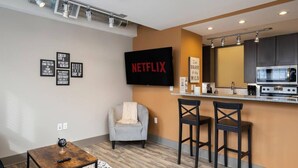 Un salon moderne avec un fauteuil gris, une table basse en bois et un coin bar équipé de deux tabourets. Un téléviseur à écran plat, fixé au mur, affiche le logo de Netflix.