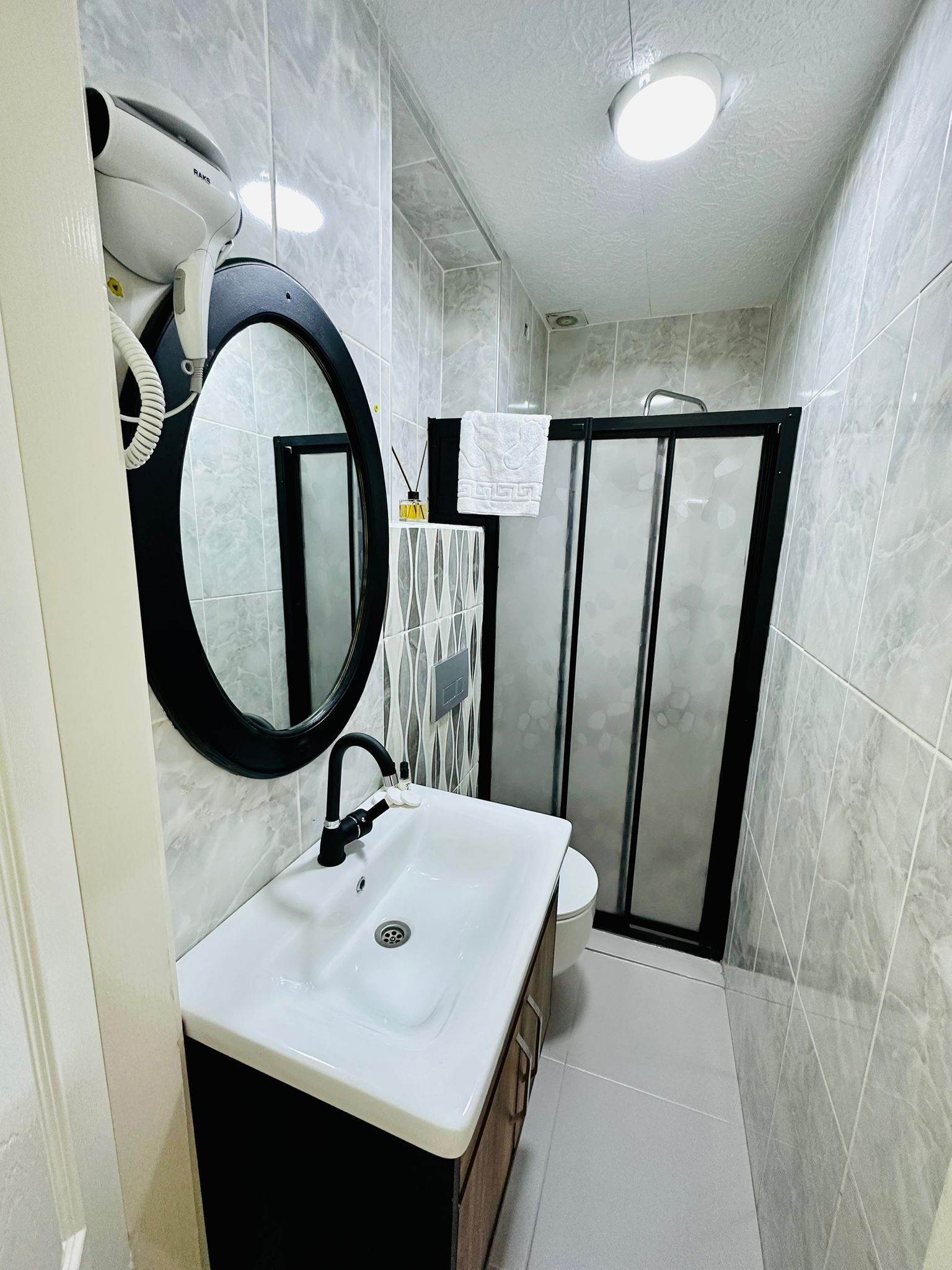 A bathroom with a white sink, black faucet, and a round mirror. There is a towel hanging on the wall and a shower with a glass door.