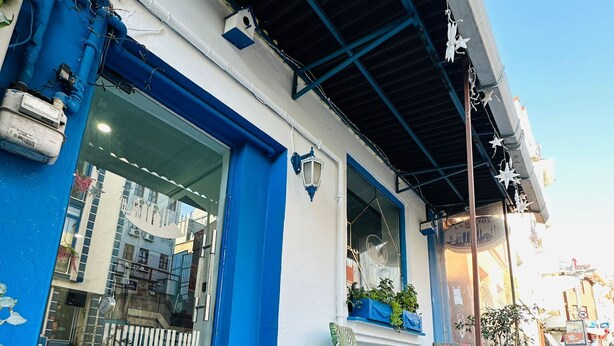 Un edificio con fachada azul y blanca, una entrada visible y un pequeño espacio al aire libre con sillas y una mesa.