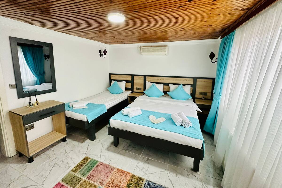 A hotel room with two single beds, a wooden ceiling, a mirror, a small bedside table, and a colorful rug.
