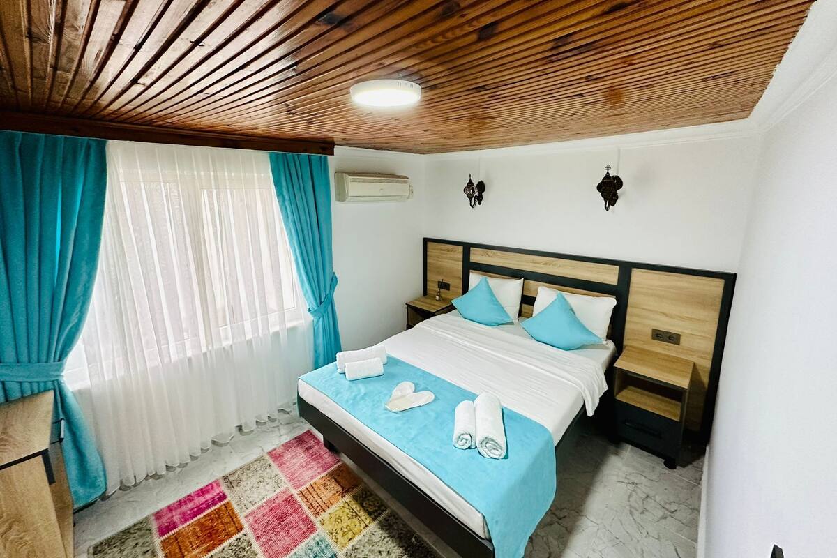 A bedroom with a wooden ceiling, a bed with blue and white bedding, a nightstand, a wall-mounted air conditioner, and a colorful rug.