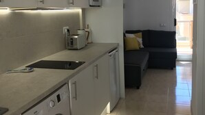 Een kleine keuken met een wasmachine, een magnetron en een broodrooster. In de aangrenzende woonkamer staat een donkere bank.