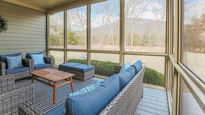 Un espacio de estar al aire libre techado con mobiliario de mimbre, una mesa de madera y vistas a un paisaje natural a través de grandes ventanales.
