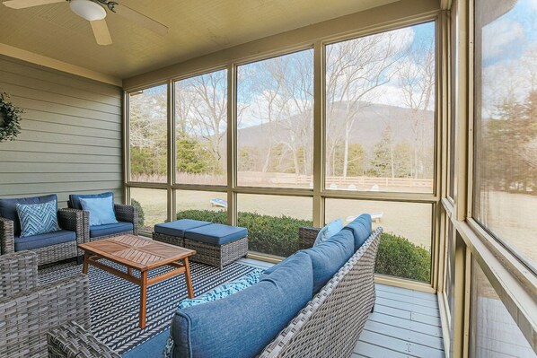 Un espace extérieur aménagé, abrité et doté de mobilier en osier, d’une table en bois et offrant une vue sur un paysage naturel grâce à de grandes fenêtres.