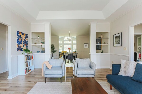 A spacious living room with a wooden floor, two grey sofas, a coffee table, and a bookshelf.