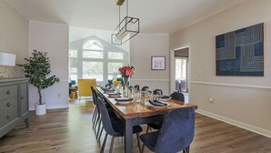 Un comedor con una mesa de madera dispuesta para seis personas, rodeada de sillas, una planta en maceta y una lámpara colgante moderna.