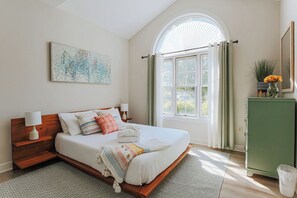 A bedroom with a large bed, a nightstand, a dresser, a window with curtains, and a painting on the wall.