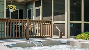 Un jacuzzi con agua corriendo, una terraza de madera y una casa con grandes ventanales y una maceta colgante.