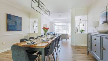 A dining area with a long wooden table set for six, flanked by chairs, a modern pendant light, and a view of a kitchen area.