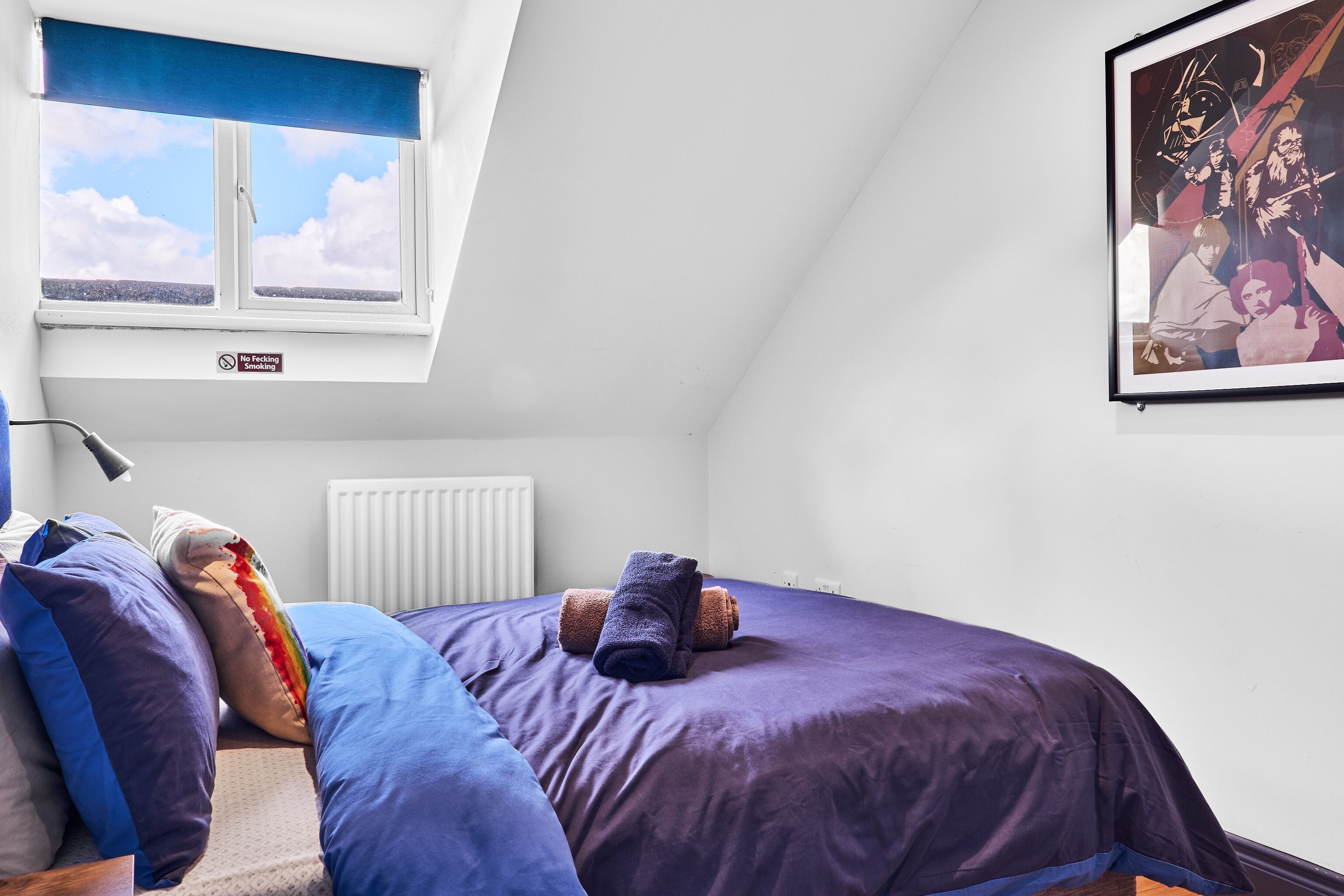 Een slaapkamer met dakraam, een bed met blauwe en paarse beddengoed, een ingelijst schilderij aan de muur, en een ‘Niet Roken’ bord.