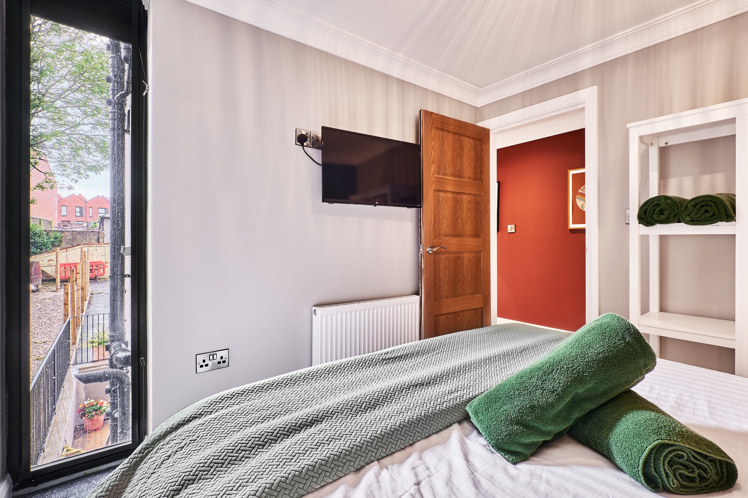Een slaapkamer met een bed, een aan de muur bevestigde televisie, een radiator en uitzicht op buiten.