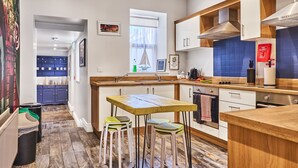A modern kitchen with a wooden table and stools, a blue tiled backsplash, and a window with sheer curtains. - Forty Four North (Londonderry)
