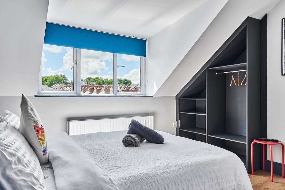 Un dormitorio con tragaluz, una cama con una toalla enrollada, un taburete rojo y un armario negro.